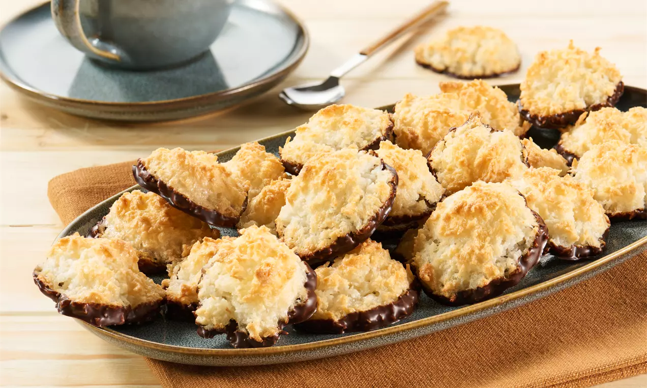 Goldbraune Kokosmakronen mit glänzend getunkter Schokoladenunterseite, locker angeordnet auf einer dunkelgrauen Platte mit orangem Tuch darunter