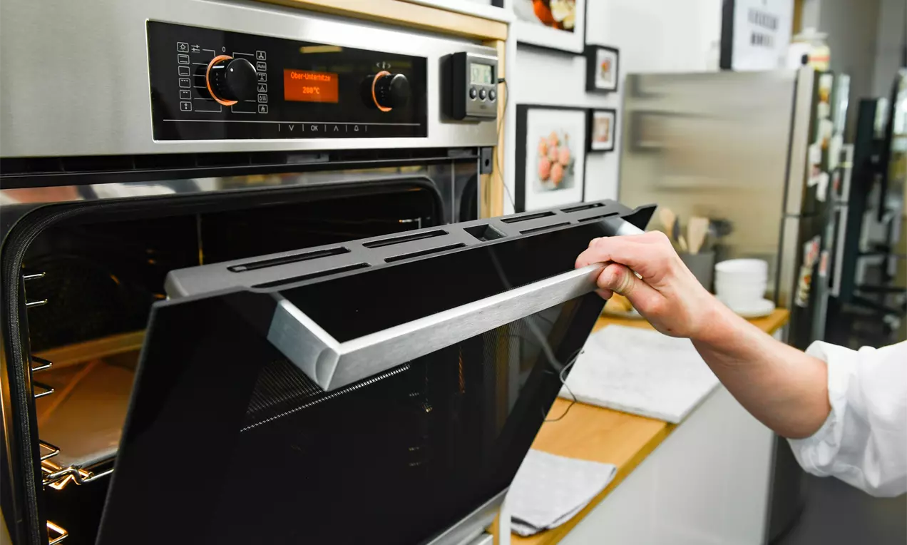 Eine geöffnete Edelstahlbackofentür mit schwarzer Front, gehalten von einer Hand, im modern eingerichteten Küchenumfeld