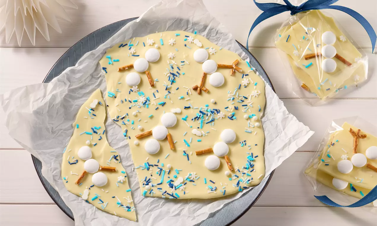 Bruchschokolade aus weißer Schokolade, geschmückt mit blauen Streuseln, weißen Schokomünzen und Brezeln, auf Backpapier angerichtet