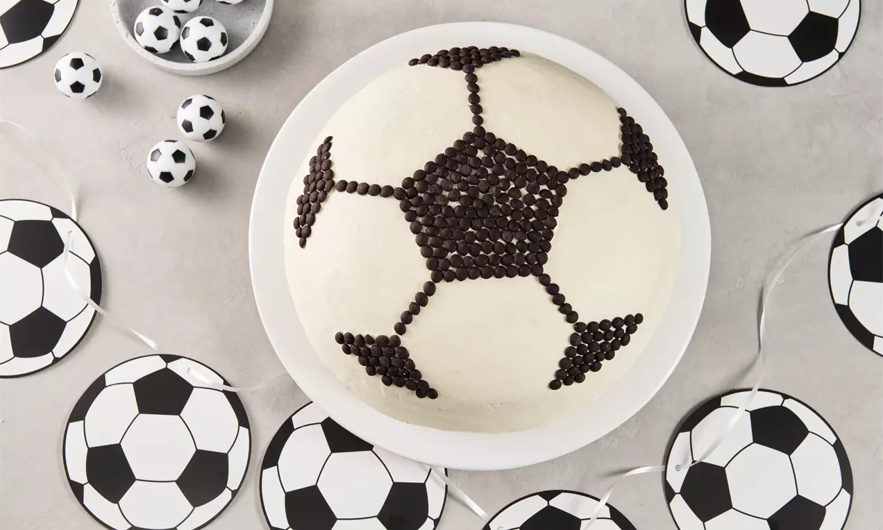 Runde Torte mit weißer Creme, verziert mit dunklen Schoko-Stückchen im Fußballmuster, auf weißem Teller, umgeben von Fußball-Dekoration