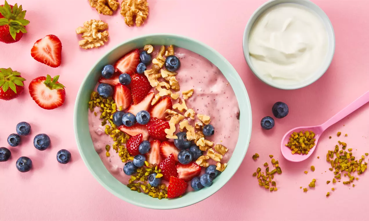 Eine rosa Smoothie-Bowl mit frischen Erdbeeren, Blaubeeren, Walnüssen und gehackten Pistazien, daneben Joghurt und ein pinker Löffel