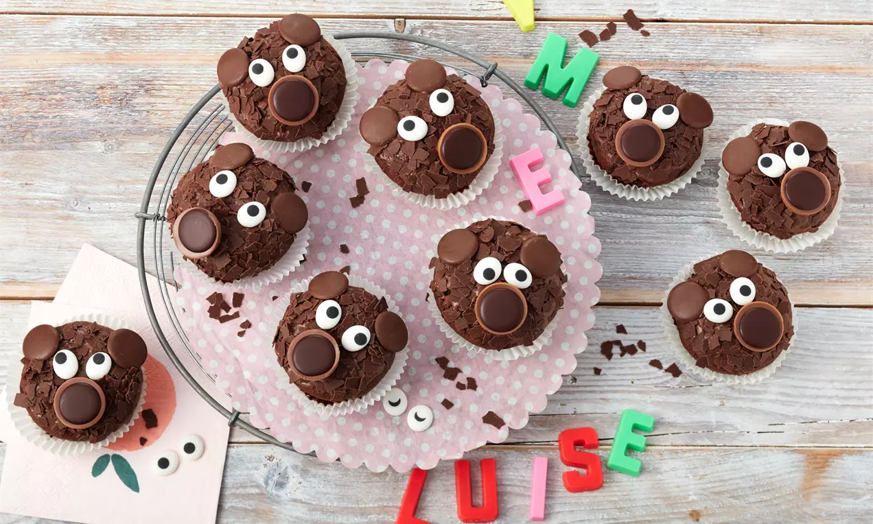 Schokoladenmuffins mit zerbröselter Schokoglasur, verziert mit Augen aus Zuckerdekor und Bärenohren aus runden Schokotalern auf rosa Papierplatte