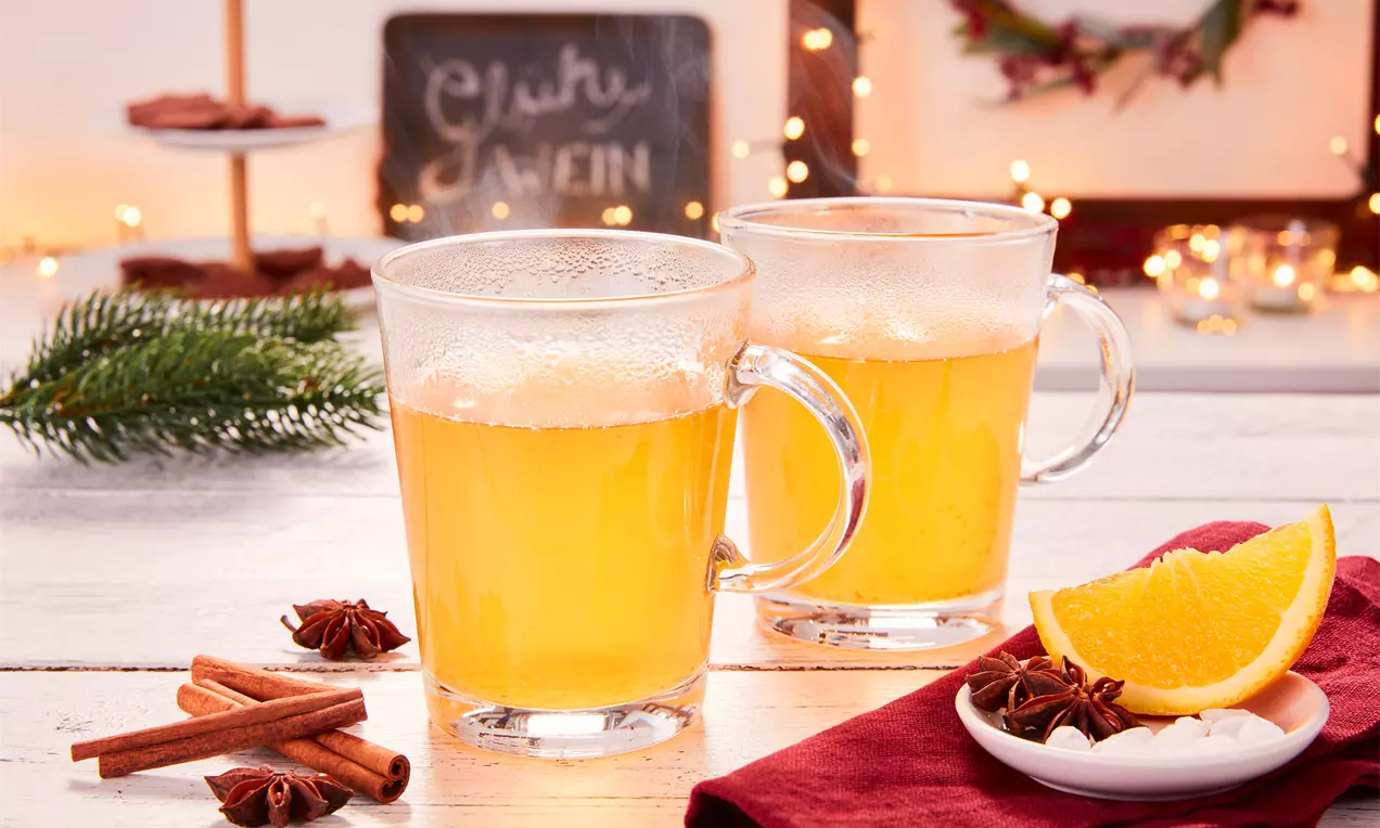 Zwei klare Glastassen mit dampfendem hellgelbem Glühwein auf weißem Holztisch, daneben Zimtstangen, Sternanis und Orangenscheiben auf rotem Tuch