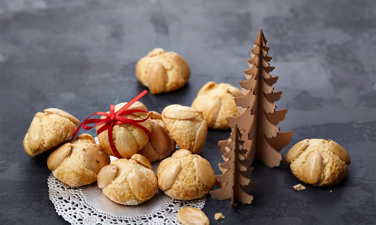 Goldbraune Bethmännchen mit leicht glänzender Oberfläche und Mandeldekor auf filigraner Papierunterlage neben brauner Tannenbaum-Dekoration