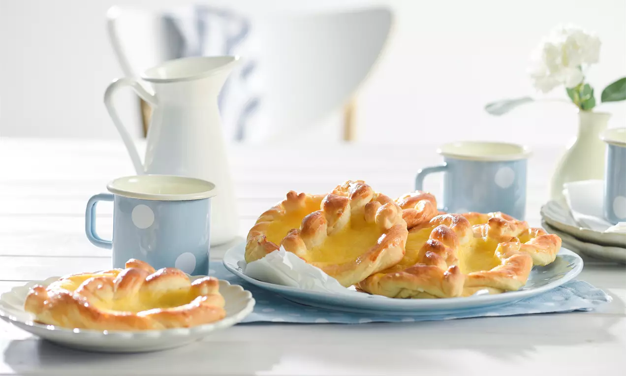 Goldbraune Puddingbrezeln mit glänzender Oberfläche und gelber Füllung auf einem weißen Teller, arrangiert neben blauen Tassen mit Punkten