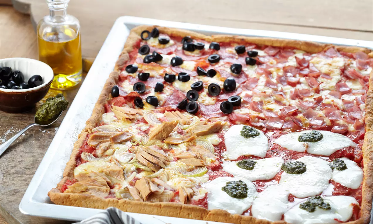 Pizza mit goldbraunem Teig auf einem Backblech, belegt mit Mozzarella, Pesto, Oliven, Thunfisch, Lauch und Schinkenwürfeln