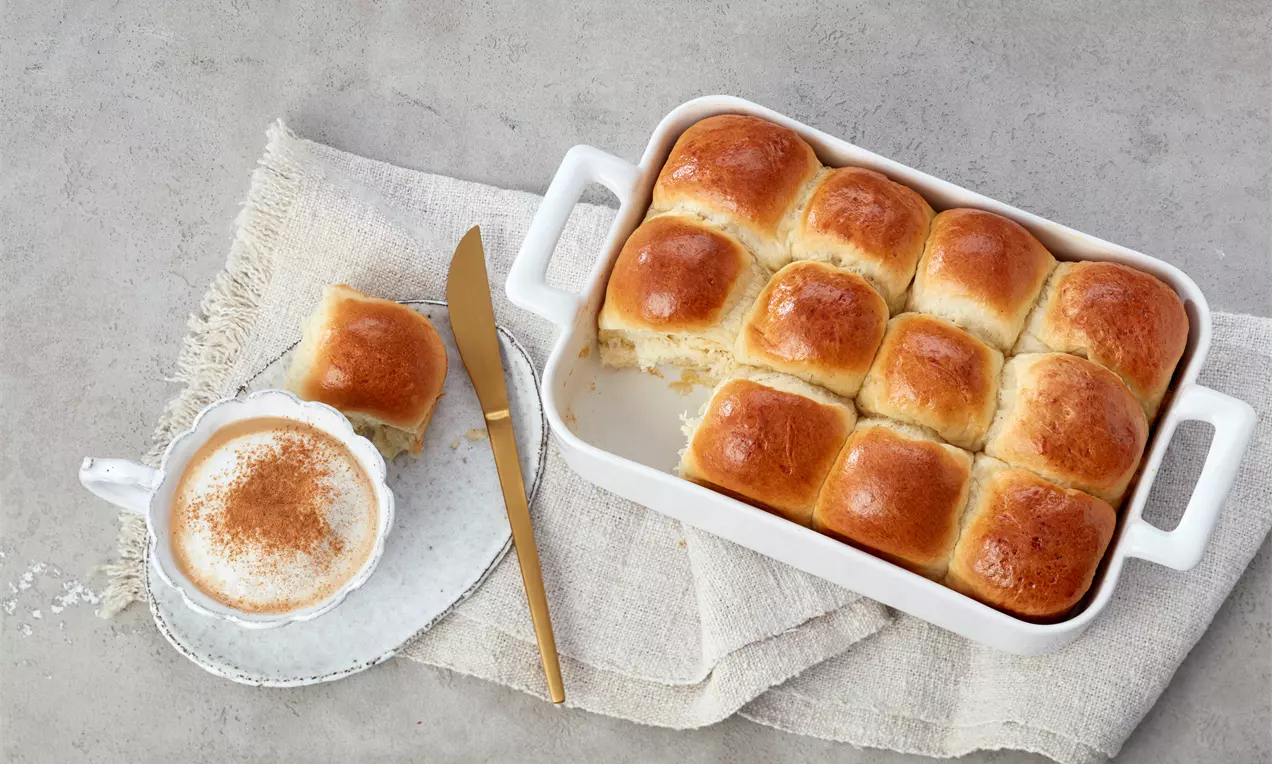 Goldbraune Milchbrötchen in einer weißen Keramikform, daneben eine Tasse mit Milchschaum und Zimt auf einem Teller mit goldfarbenem Messer