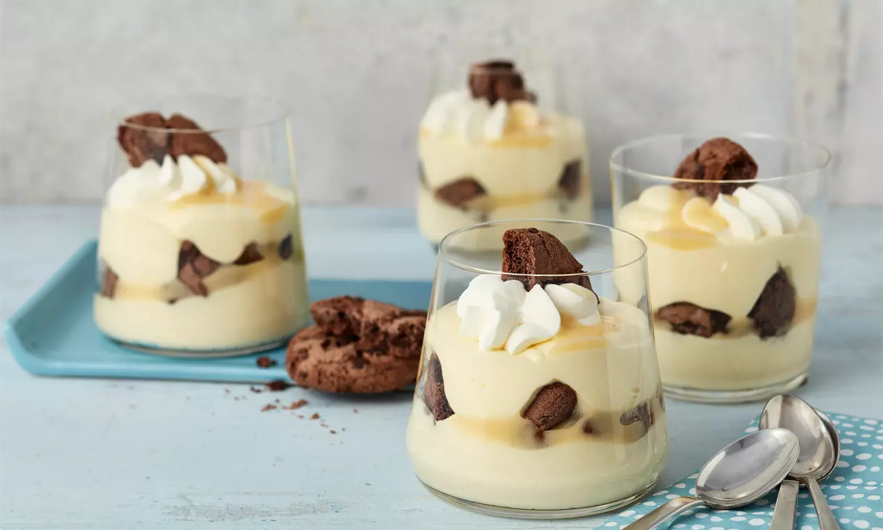 Cremige Eierlikördesserts in Gläsern mit Sahnetupfen, Schokostückchen und weicher Textur auf einem hellblauen Tisch arrangiert, AI-labeled image