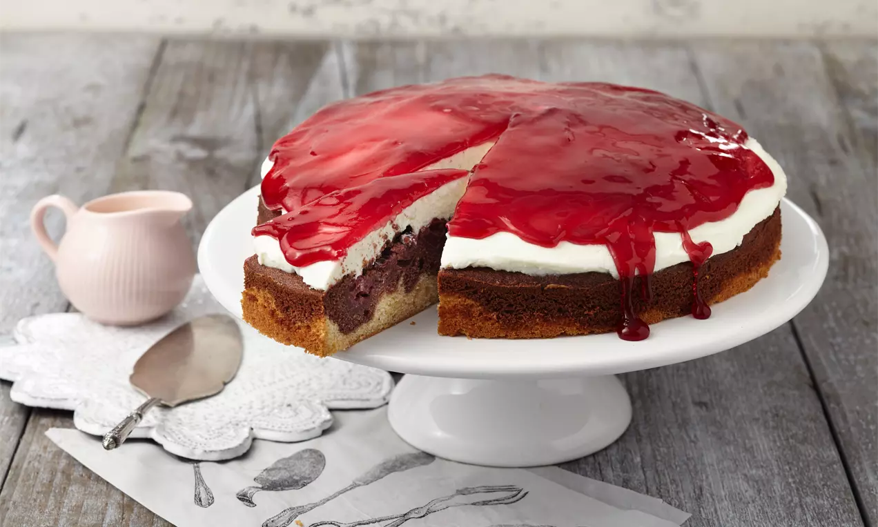 Rotkäppchen-Kuchen mit dunkler Schicht, heller Creme und glänzend rotem Fruchtbelag, auf weißer Tortenplatte auf Holztisch drapiert