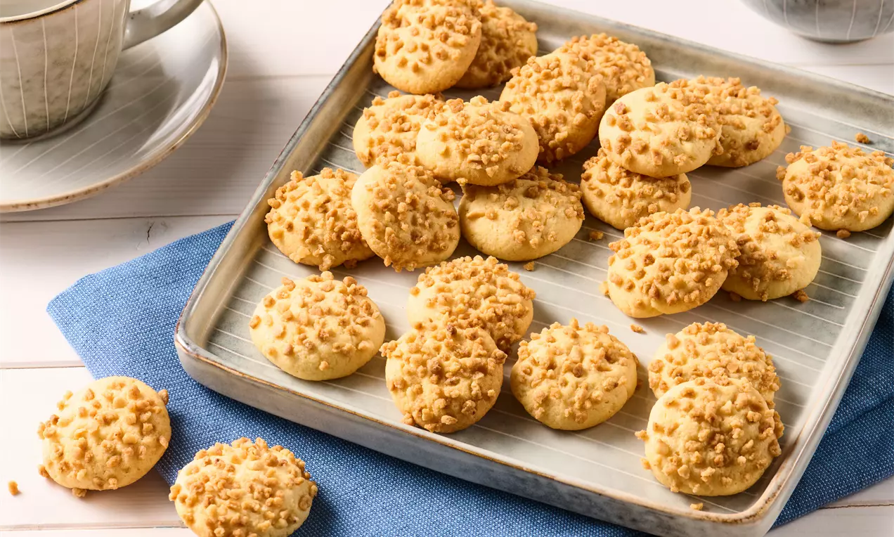 Goldgelbe Marzipanplätzchen mit gehackten Nüssen bestreut, auf einem silbernen Tablett auf blauer Stoffserviette angerichtet