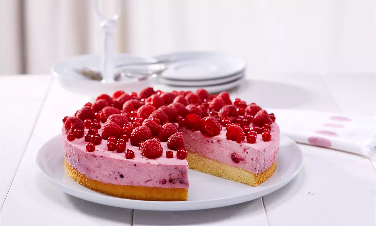 Runde Torte mit hellrosa Beerencreme auf hellem Biskuitboden, belegt mit glänzenden Himbeeren und Johannisbeeren auf weißem Teller