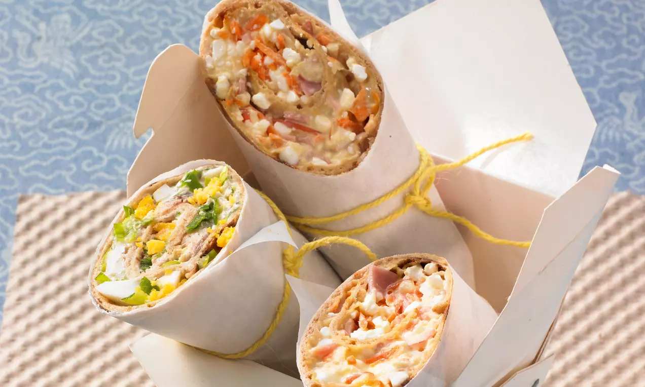 Drei helle Wraps mit bunter Füllung aus Gemüse, Salat und Fleisch, halbiert und in weißes Papier gewickelt, mit gelbem Band gebunden in einer Box