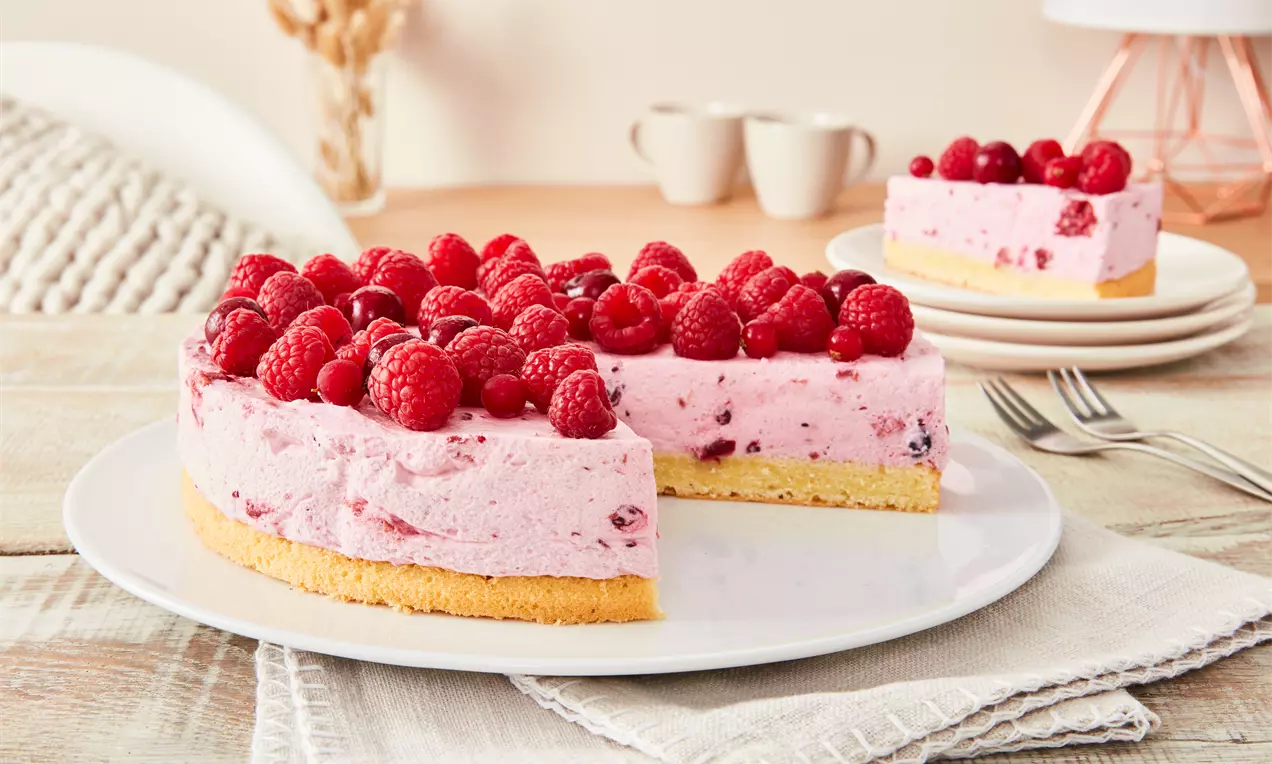 Rote-Grütze-Torte mit flaumiger rosa Füllung auf hellem Keksboden, dicht mit Himbeeren bedeckt, auf weißem Teller mit Tischdeko arrangiert