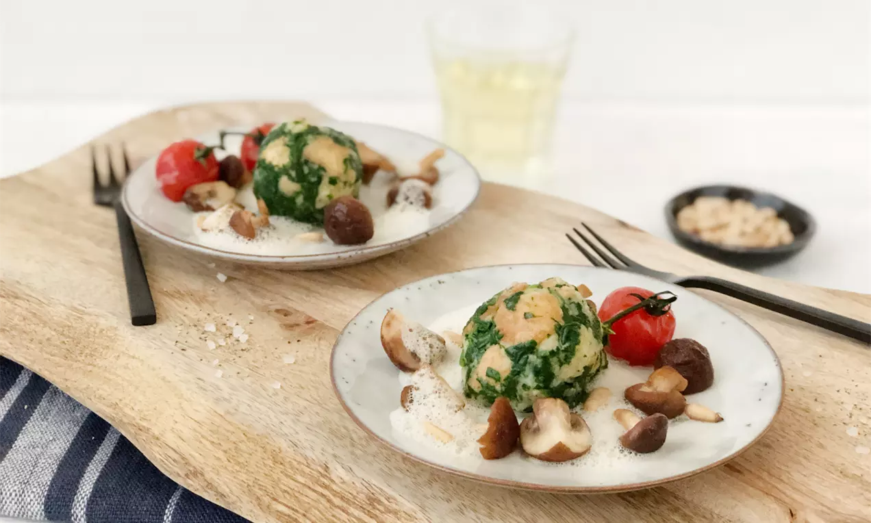 Runde Spinatknödel mit grünem Muster, angerichtet auf cremigem weißem Tomaten-Schaum, begleitet von braunen Pilzen und einer roten Kirschtomate