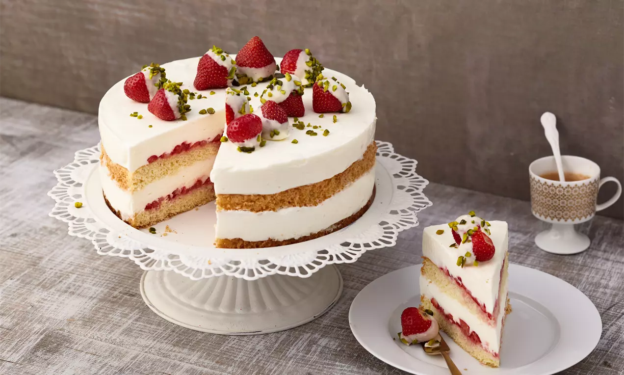 Joghurt-Frischkäse-Torte mit luftiger weißer Creme, Erdbeeren und Pistazien dekoriert auf einer filigranen Tortenplatte aus Porzellan