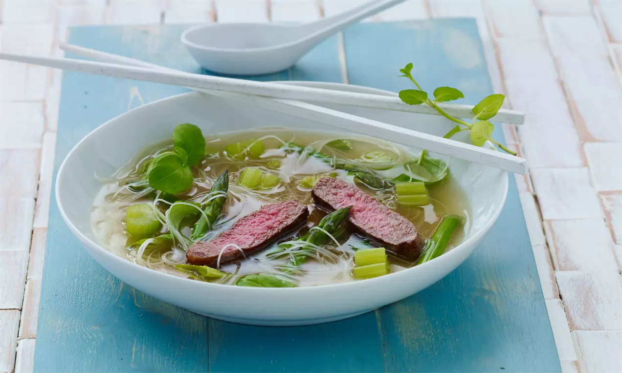 Asiatische Nudelsuppe in einer weißen Schale mit klarer Brühe, zarten Reisnudeln, grünem Gemüse und zwei rosa Rindfleischscheiben