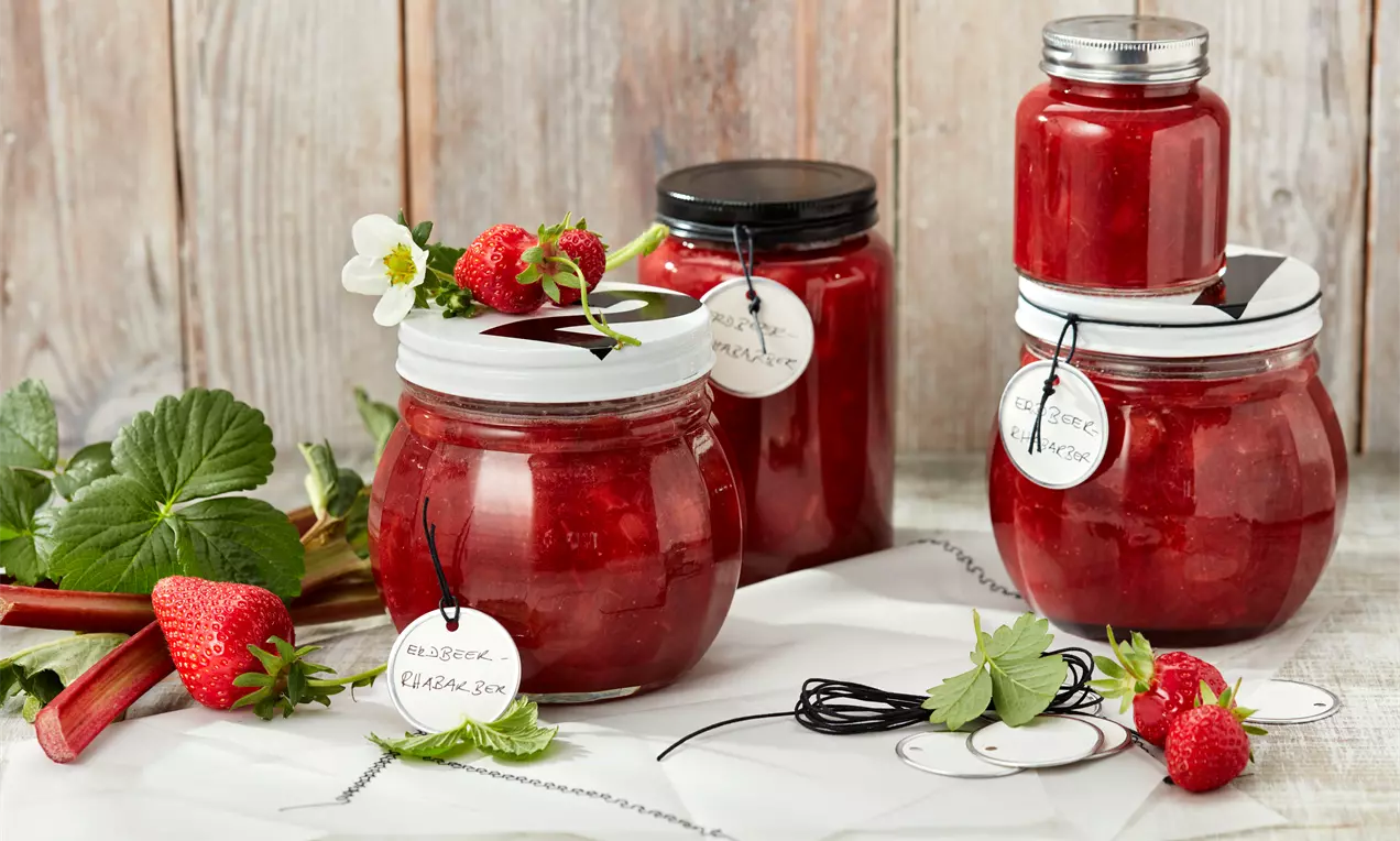 Erdbeer-Rhabarber-Konfitüre in roten, glänzenden Gläsern mit weißen Etiketten, dekoriert mit frischen Erdbeeren, Blüten und grünen Blättern