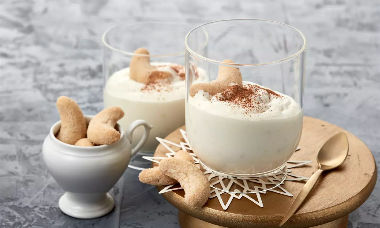 Vanillefarbene, cremige Dessertfüllung in Glas serviert, mit braunem Zimt bestäubt und von goldbraunen Kipferln garniert auf Holzplatte