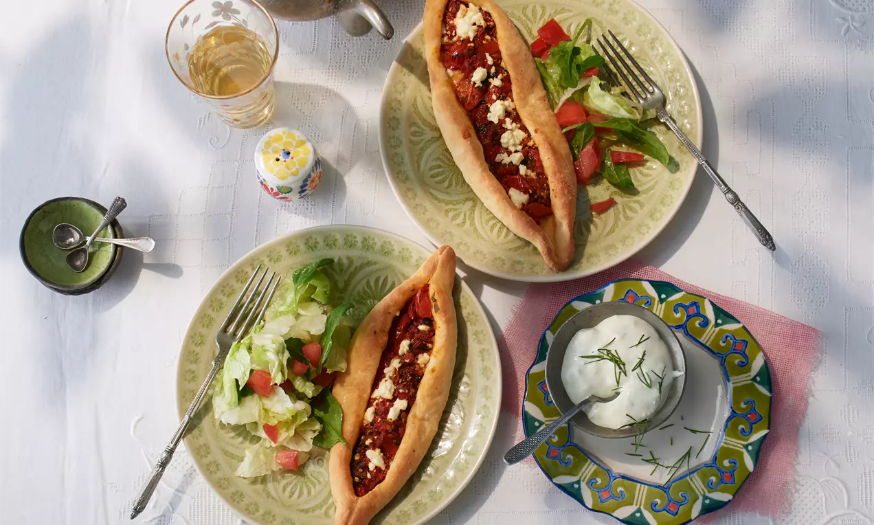 Vegetarische Pide mit goldbraunem Teig, würziger Füllung und Feta auf einem hellgrünen Teller, dazu frischer Salat und Joghurt-Dip