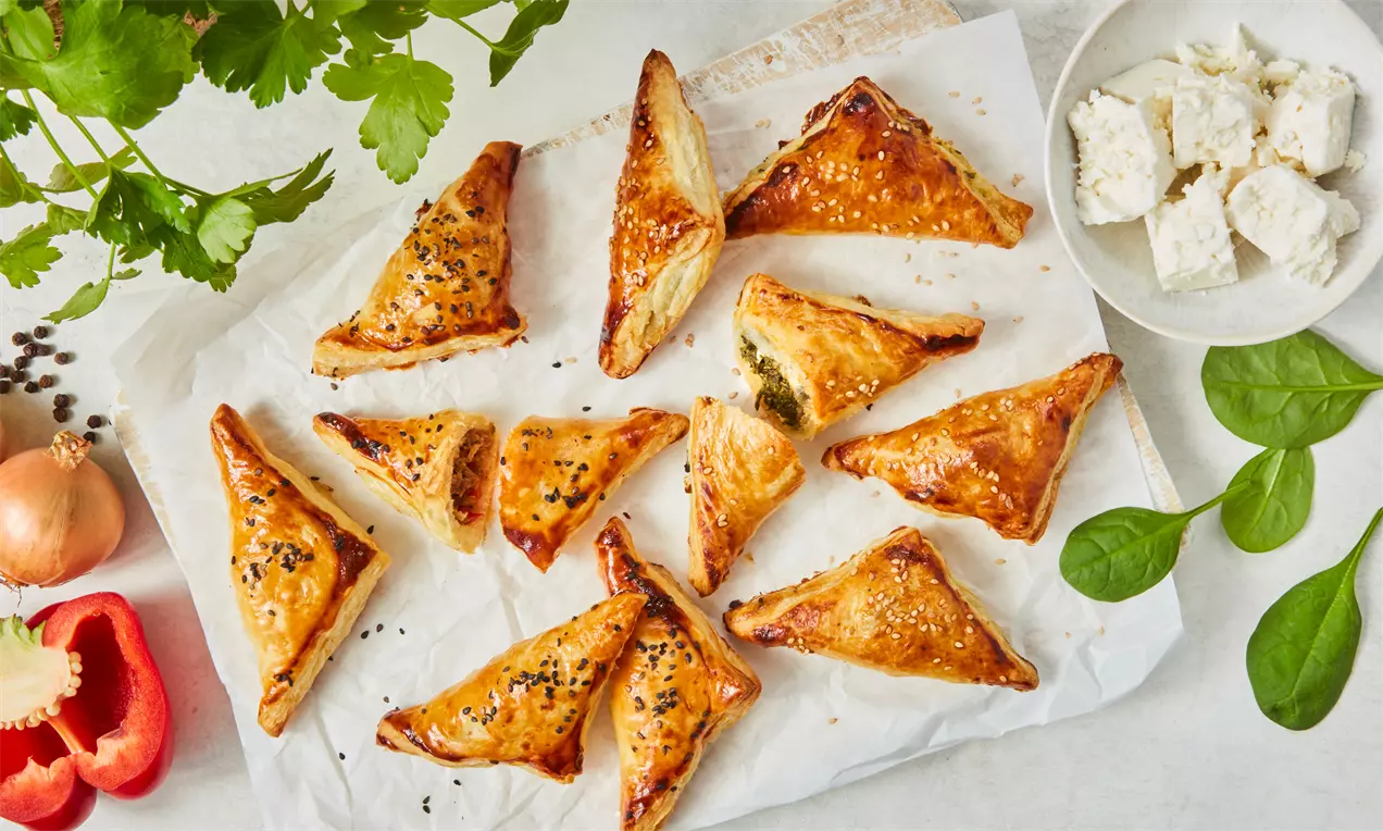 Goldbraune, dreieckige Blätterteigtaschen mit schwarzem Sesam auf Backpapier, daneben Feta, Babyspinat und frisches Gemüse