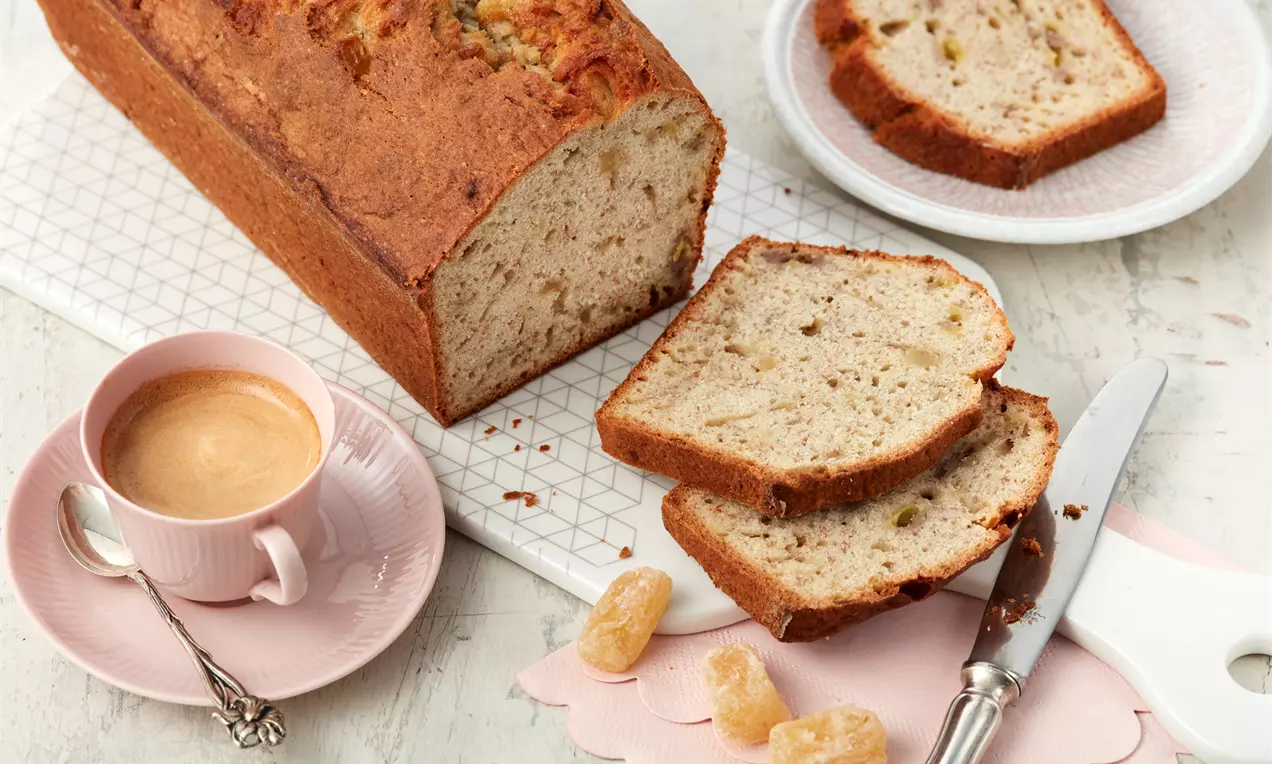 Ein goldbraunes Bananenbrot mit gesprenkelter, saftiger Krume, serviert in Scheiben neben Kandis, rosa Tasse und Messer auf einem weißen Brett