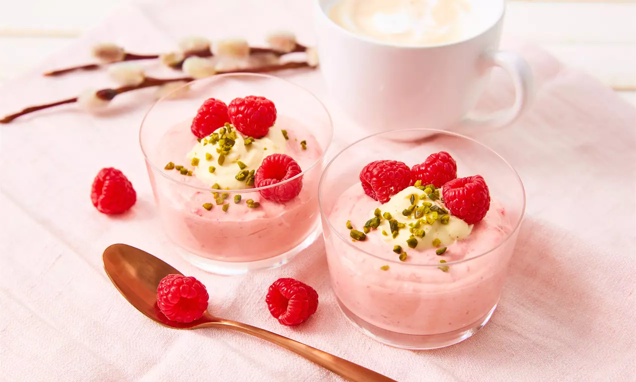 Zwei Gläser mit zartrosa Himbeer-Mousse, garniert mit Sahnehäubchen, frischen Himbeeren und gehackten Pistazien auf rosafarbenem Tuch.