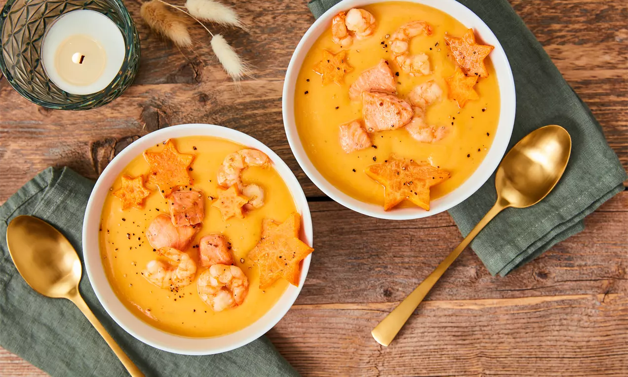 Cremige orangefarbene Süßkartoffelsuppe in weißen Schalen, garniert mit rosa Garnelen, lachsfarbenen Stücken und sternförmigen Chips