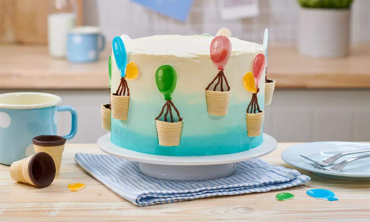 Runde Torte mit weiß-blauem Farbverlauf, verziert mit bunten Luftballons aus Schokoladenkörbchen und Zuckerdeko, auf weißer Tortenplatte