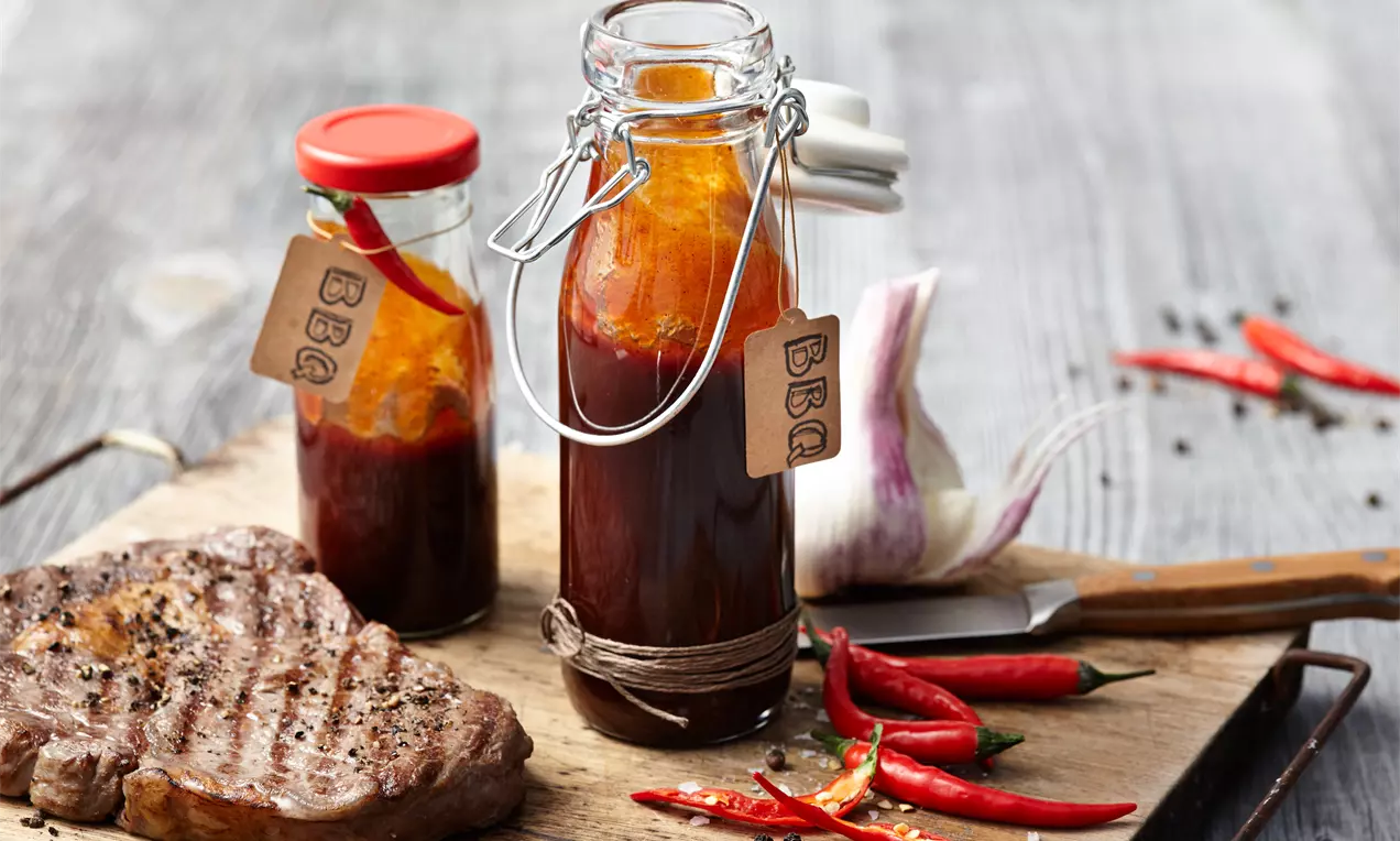 Barbecue-Soße in Glasflaschen mit braun-rötlichem Inhalt, neben Grillfleisch, roten Chilischoten und Knoblauch auf Holzbrett drapiert