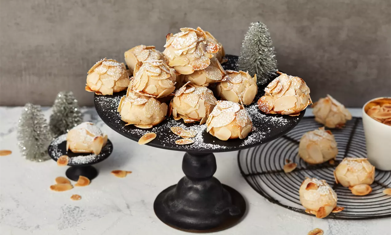 Goldbraune Marzipanhäufchen mit knusprigen Mandelblättchen und Puderzucker auf schwarzer Etagere, dekoriert mit kleinen Tannenbäumchen