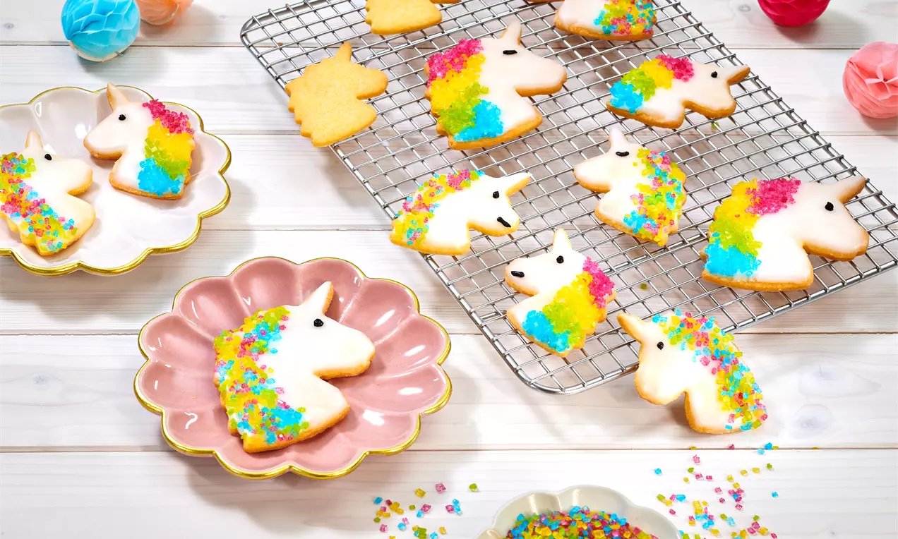 Einhorn-Kekse mit bunter Zuckerdeko auf weißer Glasur, gelegt auf rosa und weißen Tellern sowie einem Gitter, umgeben von Streusel und Deko