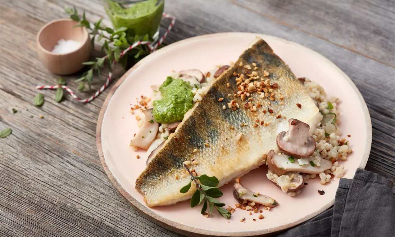 Goldbraun gebratenes Zanderfilet auf cremigem Pilzrisotto mit grünem Feldsalat-Pesto, dekoriert auf einem weißen Teller mit Holzuntergrund