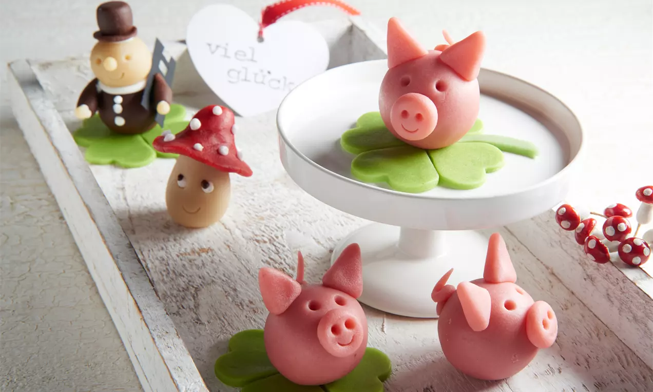 Rosa Marzipan-Glücksschweinchen mit runden Körpern und detaillierten Gesichtszügen, auf grünen Kleeblättern und einer weißen Dekoplatte arrangiert