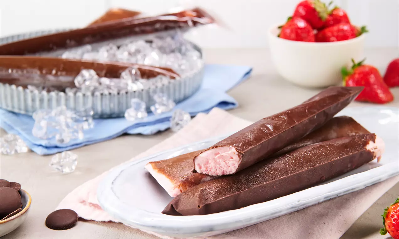 Schokoladenüberzogene Eis-Pops mit rosafarbener Füllung auf einem weißen Teller, im Hintergrund Erdbeeren und Eiswürfel auf blauem Tuch