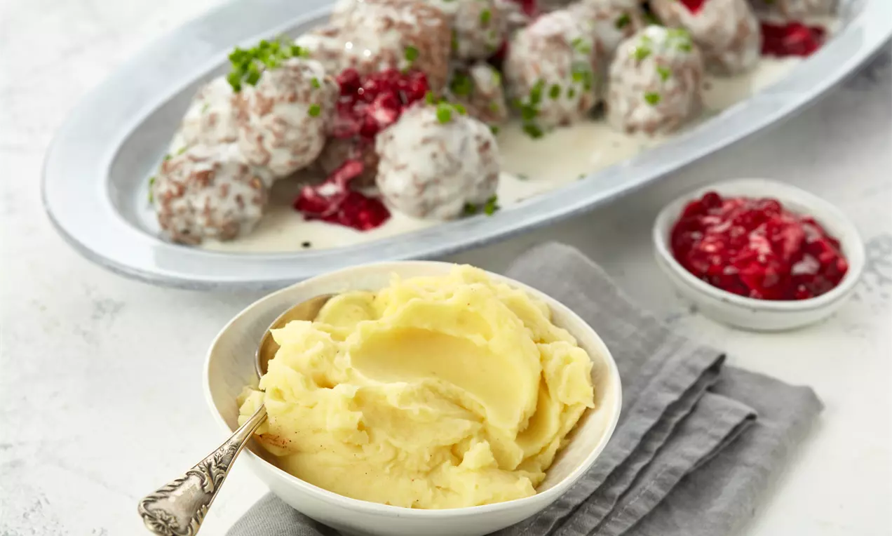 Goldgelbes, cremiges Kartoffelpüree in einer weißen Schale mit silbernem Löffel vor Köttbullar in Sahnesauce und roten Preiselbeeren auf ovaler Platte