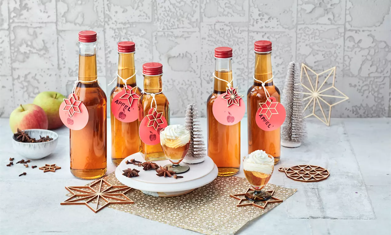 Mehrere schlanke Glasflaschen mit goldbraunem Likör und roten Verschlüssen, dekoriert mit roten Anhängern, auf hellem Untergrund mit weihnachtlichem Schmuck