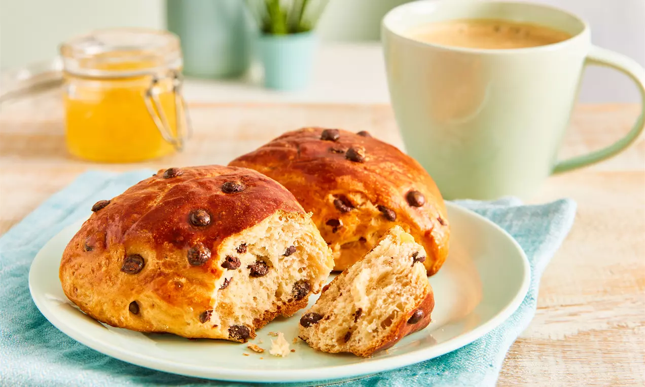 Zwei goldbraune Schokobrötchen mit glänzender Oberfläche und dunklen Schokostückchen auf einem Teller, daneben eine Tasse Kaffee