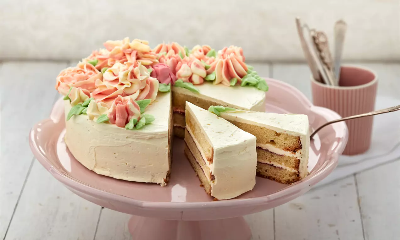 Torte mit buttercremeglatter Oberfläche und pastellfarbenen Blüten aus Sahne, auf rosa Tortenplatte mit angeschnittenem Stück und Besteck im Hintergrund