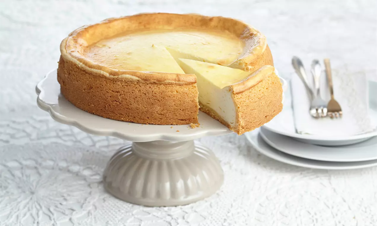 Käsekuchen mit Puddingpulver mit cremig hellgelber Füllung, goldbrauner Mürbeteigkruste und leichtem Puderzucker auf weißem Teller
