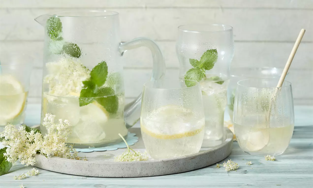 Holunderblüten-Limonade in gläsernem Krug und Gläsern mit Zitronenscheiben, Minzblättern und Blüten auf heller Steinplatte angerichtet