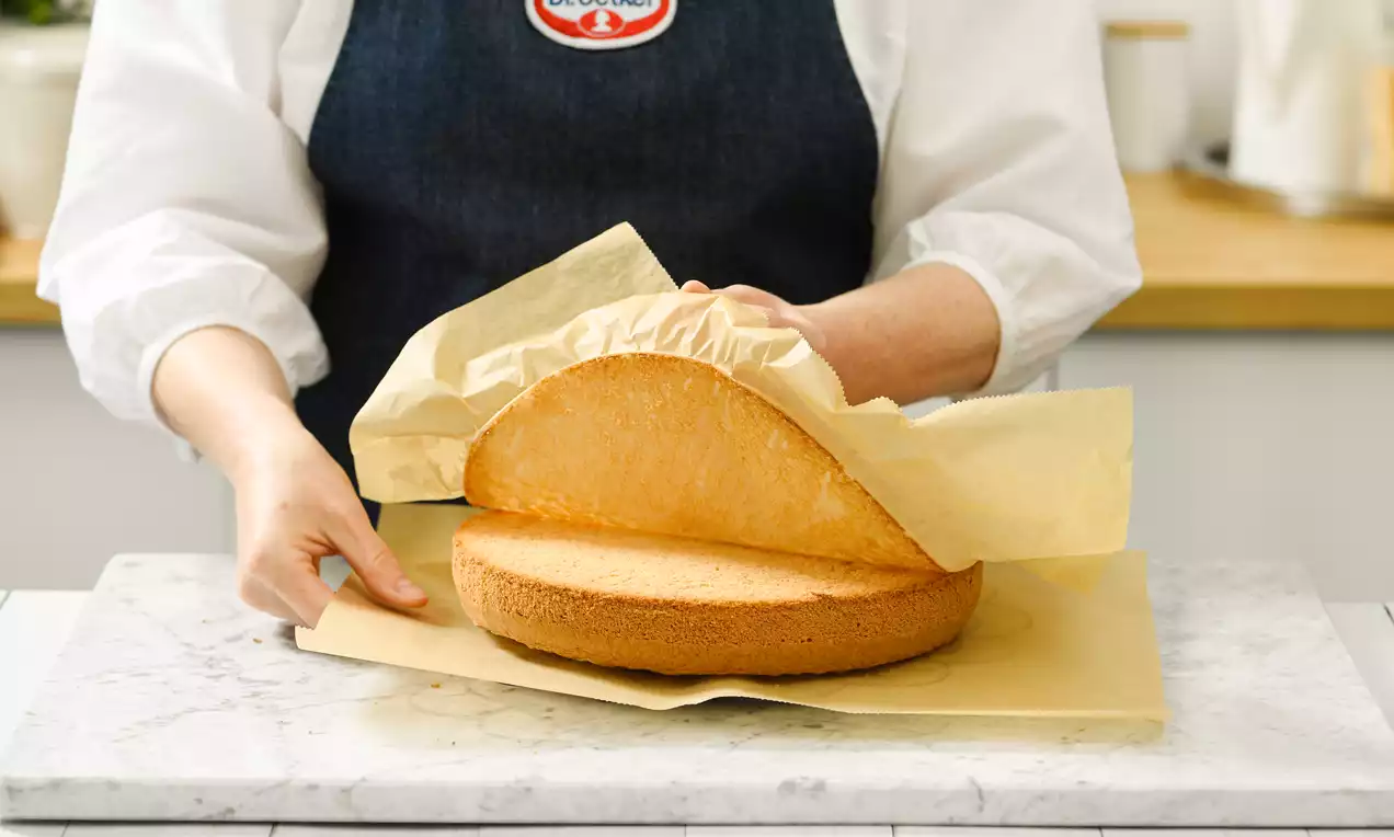 Goldgelber, runder Biskuit auf hellem Backpapier, eine Hand hebt vorsichtig das leicht gewellte Pergament von der weichen Oberfläche ab