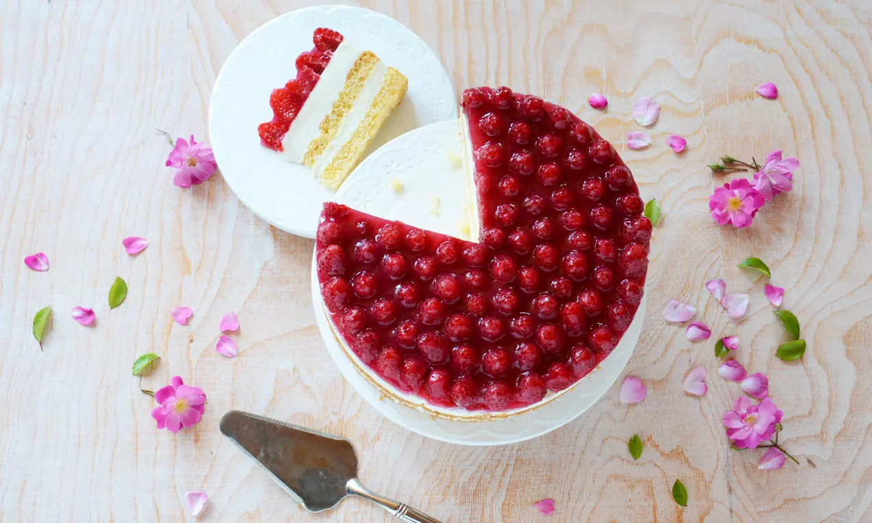Himbeer-Käse-Sahnetorte mit leuchtend roten Himbeeren und cremiger Füllung auf weißer Tortenplatte, umgeben von pinken Blumenblüten