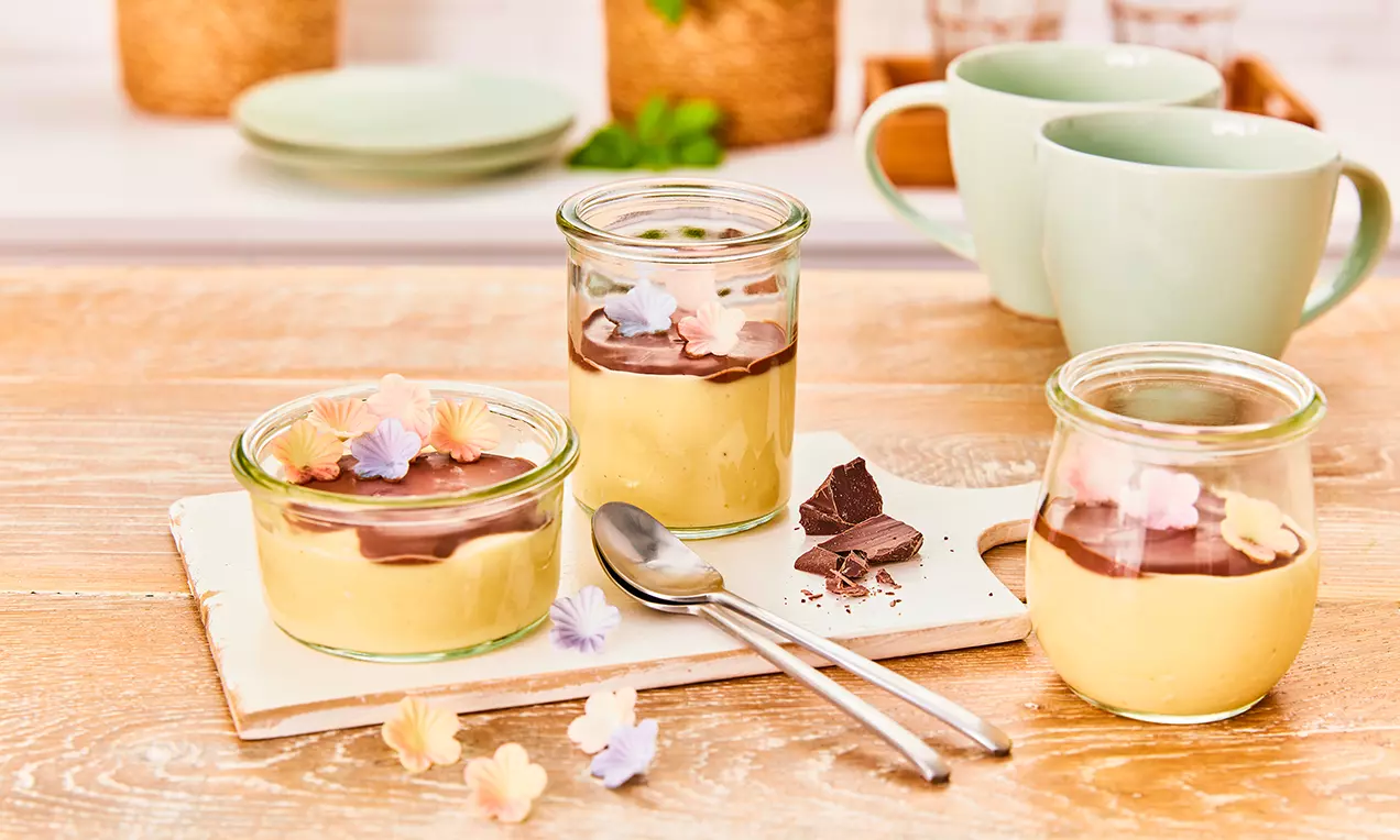 Veganer Vanillepudding in Gläsern mit einer glänzenden dunkelbraunen Schokosoße, dekoriert mit essbaren Blüten auf einem weißen Brett