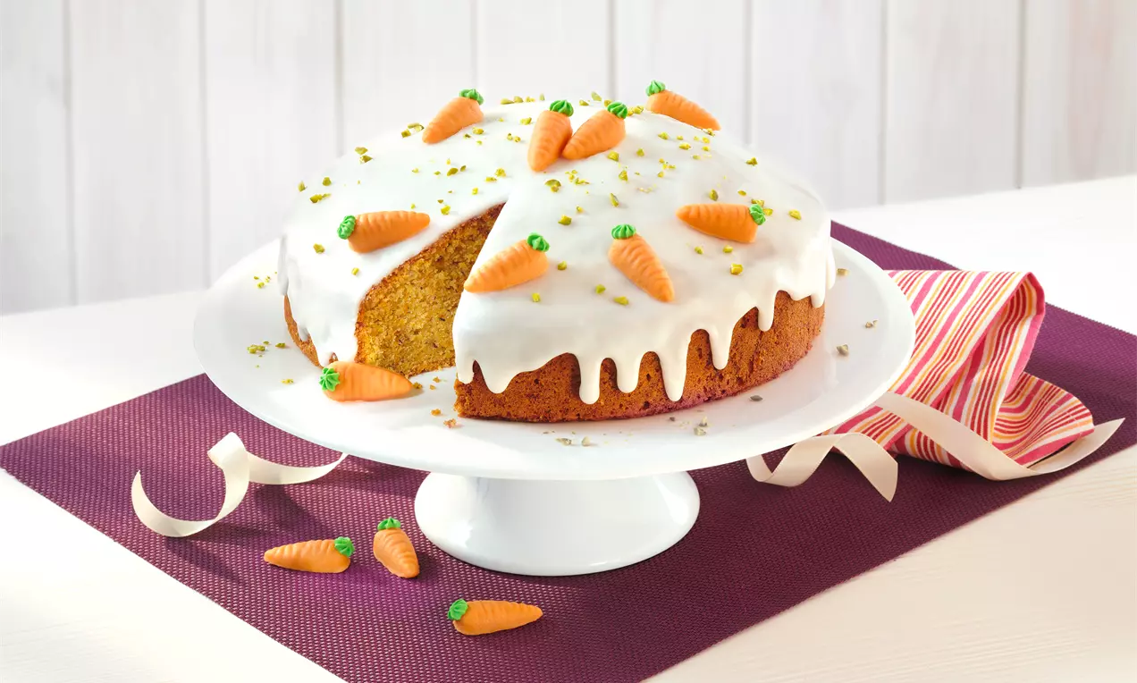 Rüblikuchen mit weißem Zuckerguss, dekoriert mit orangefarbenen Marzipan-Rübli und grünen Pistazien, auf weißer Tortenplatte serviert