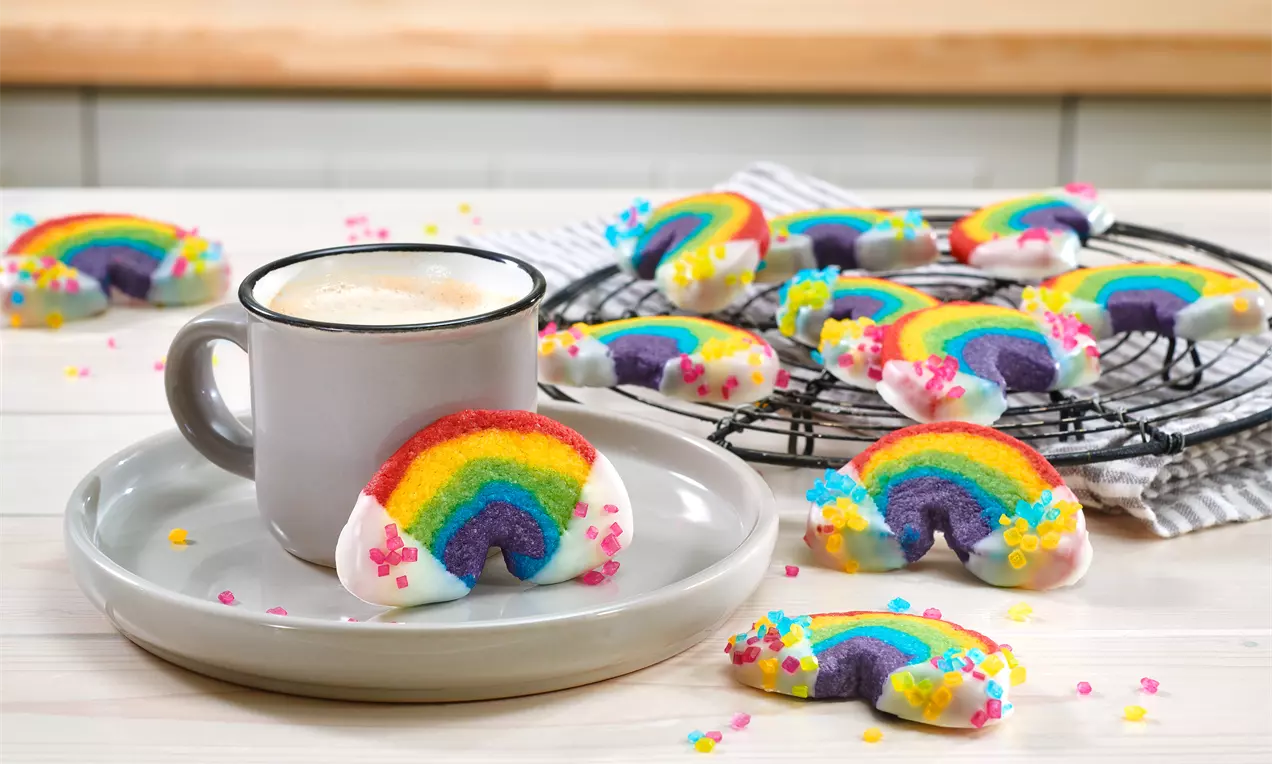 Halbkreisförmige Regenbogenkekse mit bunten Streifen, weißer Glasur und bunten Zuckerperlen auf Teller und Kuchengitter angerichtet