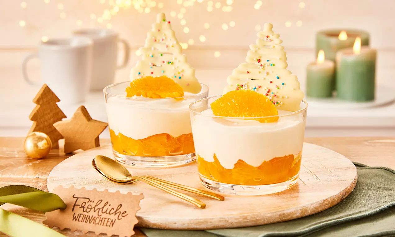 Weiße Schokoladenmousse in einem Glas auf leuchtend orangefarbenem Orangenkompott, dekoriert mit einer Orangenfiletscheibe und einem weißen Schokoladentannenbaum