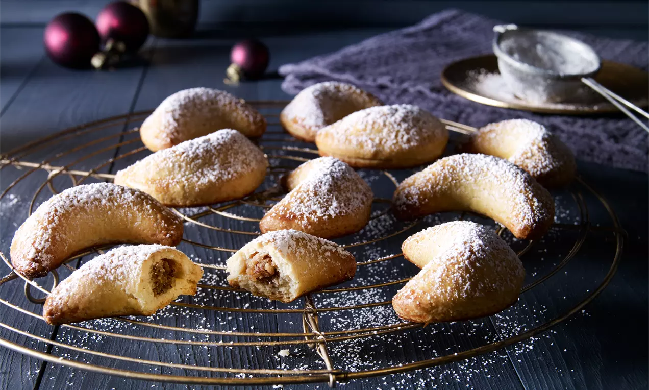 Goldgelbe Kipferl mit gebogener Form, gefüllt mit Nuss-Marzipan, bestäubt mit Puderzucker auf einem goldenen Gitter mit dunkler Dekoration