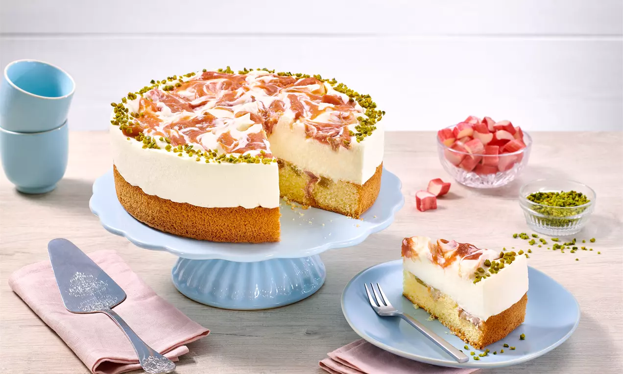 Rhabarber-Frischkäse-Kuchen mit weißer Cremefüllung, zartem gelbem Boden, Rhabarberstücken und Pistazienringen auf einer blauen Tortenplatte