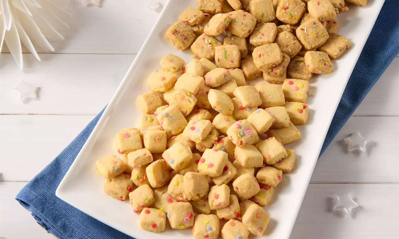 Kleine, würfelförmige Shortbread-Stücke in hellem Goldgelb mit bunten Streuseln auf einer länglichen weißen Platte serviert