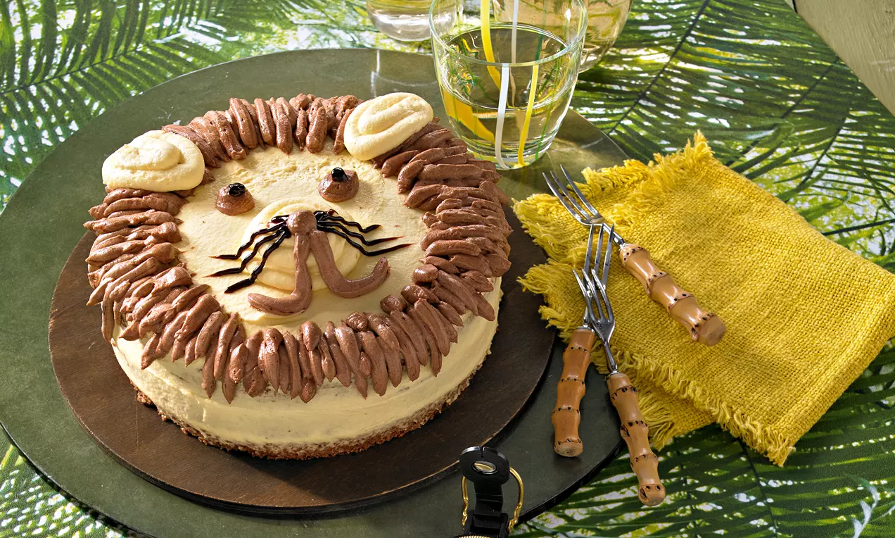 Runde Torte mit brauner Keksbasis, heller Creme, Schokosahne-Mähne, Gesicht aus Zuckerguss, Ohren aus Keksen auf grünem Dschungel-Setting
